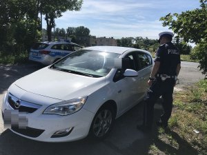 Zdjęcie przedstawia umundurowanego policjanta ruchu drogowego prowadzącego kontrolę stojącego obok niego auta osobowego.