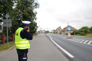 Zdjęcie przedstawia umundurowanego policjanta ruchu drogowego prowadzącego kontrolę prędkości przemieszczających się po trasie aut.