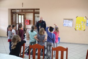 Zdjęcie przedstawia umundurowanego policjanta prowadzącego zajęcia dla stojących przed nim dzieci.