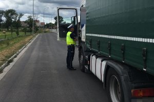 Zdjęcie przedstawia umundurowanego policjanta ruchu drogowego prowadzącego kontrolę auta ciężarowego