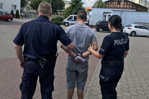 Zdjęcie przedstawia dwoje policjantów kobietę i mężczyznę prowadzących zatrzymanego podejrzanego. podejrzany założone ma na trzymane z tyłu dłonie kajdanki.