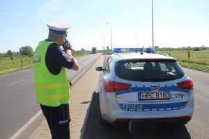 zdjęcie przedstawia policjanta ruchu drogowego prowadzącego kontrolę prędkości aut. w tle widać pojazd ciężarowy.