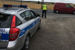 Zdjęcie przedstawia policjanta ruchu drogowego stojącego przy drodze i obserwującego ruchu pojazdów.