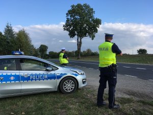 Zdjęcie przedstawia umundurowanych policjantów ruchu drogowego obserwujących ruch. obok stoi oznakowany radiowóz.