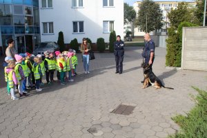 Zdjęcie przedstawia przewodnika policyjnego psa wraz z umundurowaną policjantką rozmawiających na placu komendy z dziećmi.