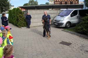 Zdjęcie przedstawia przewodnika policyjnego psa wraz z umundurowaną policjantką i policjantem rozmawiających na placu komendy z dziećmi.