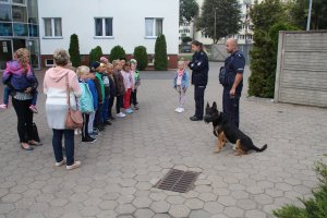 Zdjęcie przedstawia przewodnika policyjnego psa wraz z umundurowaną policjantką rozmawiających na placu komendy z dziećmi.