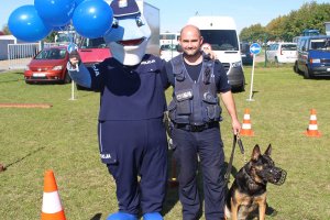 zdjęcie przedstawia maskotkę kujawsko-pomorskiej policji Polfina stojącego razem z przewodnikiem psa i psem.