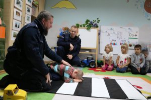 Zdjęcie przedstawia dwóch umundurowanych policjantów, z których jeden pokazuje na dziecku jak bronić się przed atakiem psa.