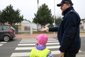 Zdjęcie przedstawia umundurowanego policjanta stojącego wspólnie z dzieckiem w rejonie przejścia dla pieszych.