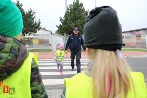 Zdjęcie przedstawia umundurowanego policjanta przechodzącego przez przejście dla pieszych razem z dzieckiem.