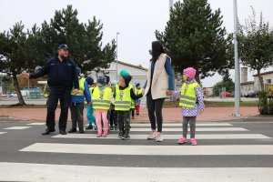 Zdjęcie przedstawia umundurowanego policjanta przechodzącego wspólnie z dziećmi i opiekunką przez przejście dla pieszych.