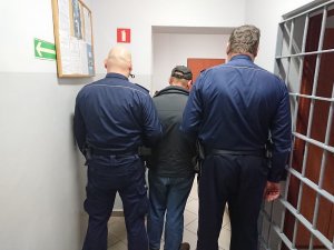 Zdjęcie przedstawia podejrzanego prowadzonego przez umundurowanych policjantów