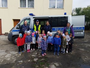 Zdjęcie przedstawia dzieci z opiekunką i policjantami stojących na tle radiowozu i pozujących do fotografii.