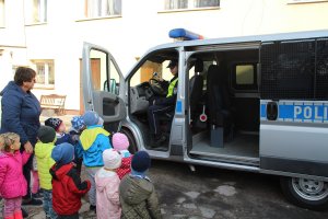 Zdjęcie przedstawia dzieci wraz z opiekunką stojących koło oznakowanego radiowozu. dzieci oglądają radiowóz. w aucie siedzi umundurowany policjant i pokazuje wyposażenie radiowozu.