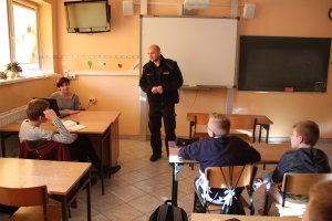 Zdjęcie przedstawia umundurowanego policjanta prowadzącego w sali lekcyjnej zajęcia dla siedzących w klasie uczniów.