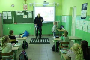 Zdjęcie przedstawia umundurowanego policjanta prowadzącego w sali lekcyjnej zajęcia dla uczniów. Policjant uczy stojącą obok niego dziewczynkę jak prawidłowo przechodzić przez przejście dla pieszych.