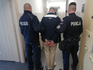 Zdjęcie przedstawia zatrzymanego prowadzonego przez dwóch umundurowanych policjantów.