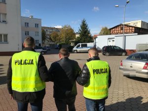 Zdjęcie przedstawia podejrzanego prowadzonego przez dwóch policjantów ubranych w kamizelki koloru żółtego z napisem policja.