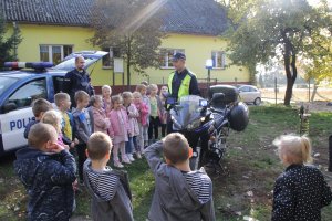 Zdjęcie przedstawia dzieci stojące obok motocykla służbowego. obok dzieci stoi dwóch umundurowanych policjantów. w tle widać oznakowany radiowóz.