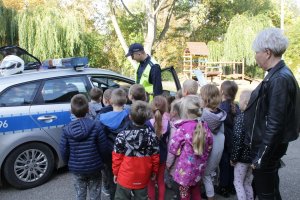 Zdjęcie przedstawia dzieci stojące obok oznakowanego radiowozu razem z policjantem i opiekunką.