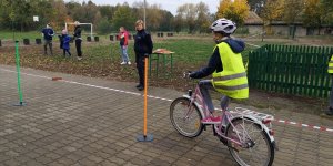 Zdjęcie przedstawia dziewczynkę egzaminowaną na kartę rowerową. w tle widać umundurowaną policjantkę oraz stojące dzieci.