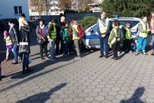 Zdjęcie przedstawia dzieci na terenie jednostki. W tle radiowóz i umundurowany policjant