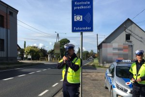 Zdjęcie przedstawia dwóch policjantów ruchu drogowego. Jeden z nich mierzy prędkość pojazdów przemieszczających się trasą, drugi stoi obok. W tle widać auta jadące drogą.