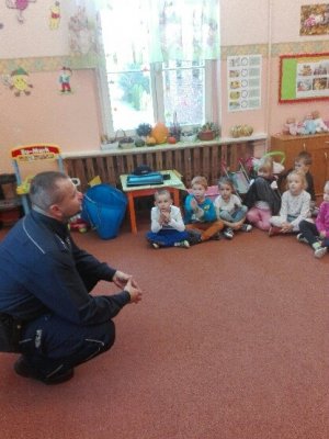 Zdjęcie przedstawia policjanta prowadzącą zajęcia dla siedzących w sali przedszkolnej dzieci. Policjant klęczy obok dzieci.