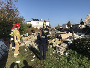 Zdjęcie przedstawia miejsce wybuchu. Na gruzowisku pracują strażacy. Obok stoi umundurowana policjantka.