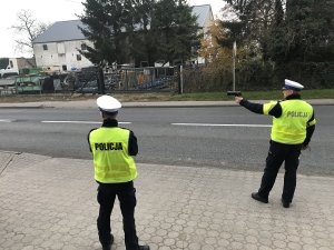 Zdjęcie przedstawia dwóch policjantów ruchu drogowego, z których jeden mierzy prędkość pojazdów.