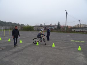 Zdjęcie przedstawia umundurowanego policjanta nadzorującego chłopca zdającego praktyczny egzamin na kartę rowerową. po lewej stronie fotografii stoi nauczyciel-mężczyzna.