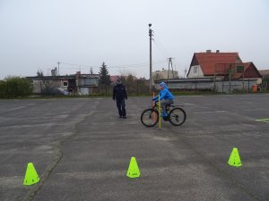 Zdjęcie przedstawia umundurowanego policjanta nadzorującego chłopca zdającego praktyczny egzamin na kartę rowerową.