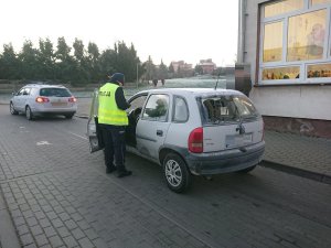 Zdjęcie przedstawia umundurowanego policjanta kontrolującego przy szkole auto osobowe