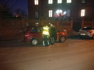 Zdjęcie przedstawia umundurowanego policjanta kontrolującego przy przedszkolu auto osobowe