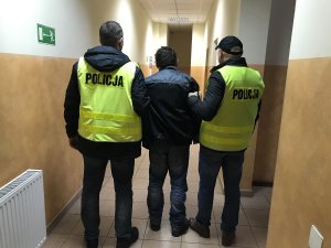 Zdjęcie przedstawia zatrzymanego podejrzanego prowadzonego przez policjantów ubranych w kamizelki z napisem policja