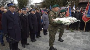 Zdjęcie przedstawia delegacje służb mundurowych składającą wiązankę kwiatów pod pomnikiem-mogiłą