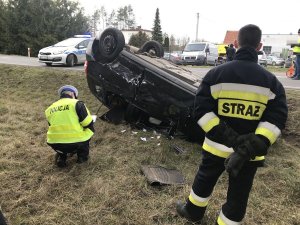 Zdjęcie przedstawia policjanta ruchu drogowego prowadzącego oględziny auta biorącego udział w wypadku. Na pierwszym lanie widać też strażaka. w tle parking z autami.