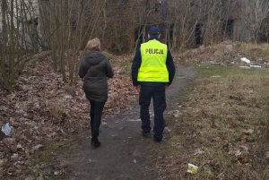 Zdjęcie przedstawia umundurowanego policjanta dodatkowo mającego ubraną kamizelkę odblaskową. Mundurowy wspólnie z pracownikiem socjalnym sprawdza miejsca przebywania bezdomnych.