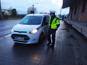 Zdjęcie przedstawia umundurowanego policjanta ruchu drogowego prowadzącego kontrolę auta osobowego