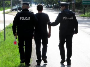 Zdjęcie przedstawia dwóch umundurowanych policjantów prowadzących zatrzymanego