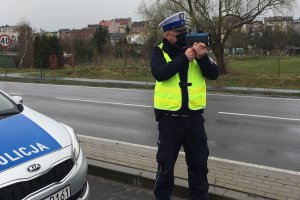 policjant mierzy prędkość aut urządzeniem. po lewej stronie zdjęcia fragment radiowozu