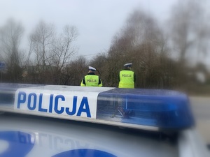 zdjęcie ilustracyjne. dach oznakowanego radiowozu. w tle stoją policjanci ubrani w kamizelki